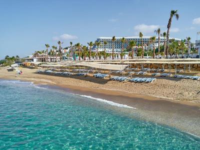 Pláž s tyrkysovou vodou a velkým hotelem v pozadí pod modrou oblohou.