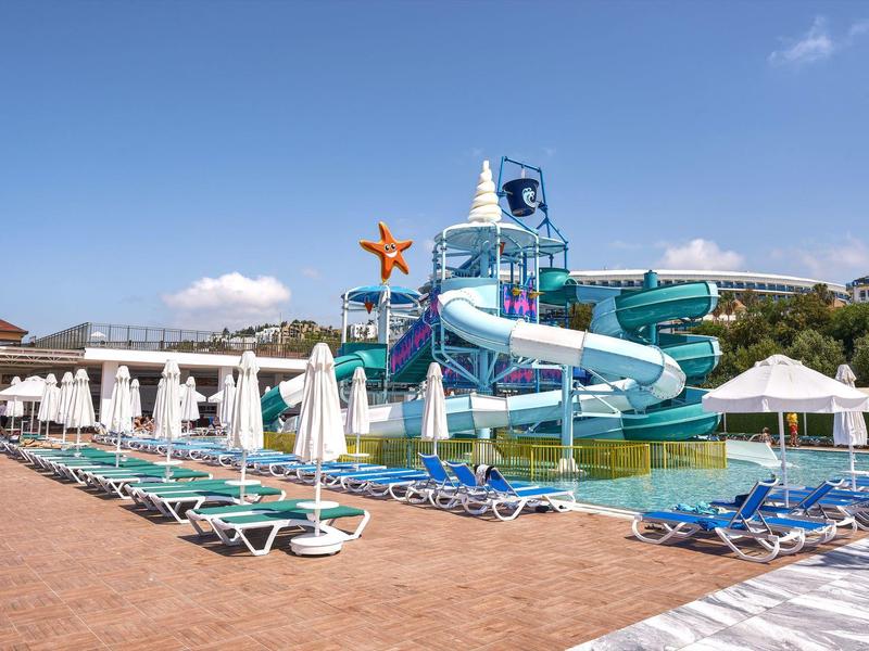 Prázdná bazénová oblast s lehátky a skluzavkami pod jasnou oblohou v rekreačním středisku.