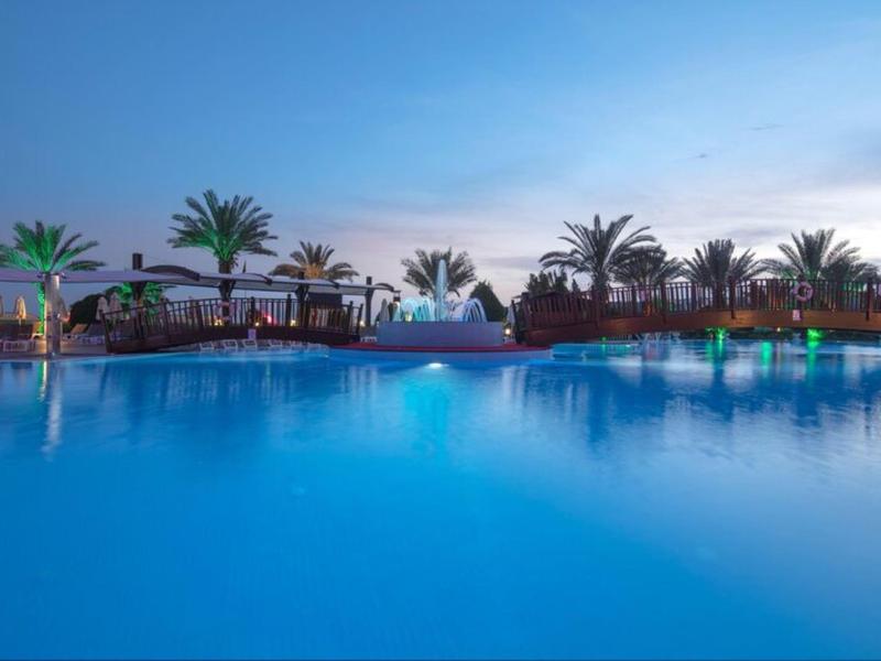 Osvětlený bazén za soumraku obklopený palmami v tropickém dovolenkovém prostředí.