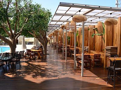 Espace extérieur d'hôtel avec terrasse couverte, tables en bois, sièges, arbres et piscine.