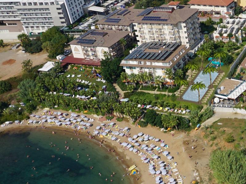 Strand mit Liegen und Sonnenschirmen, dahinter Hotels und Pool, umgeben von Grünanlagen.