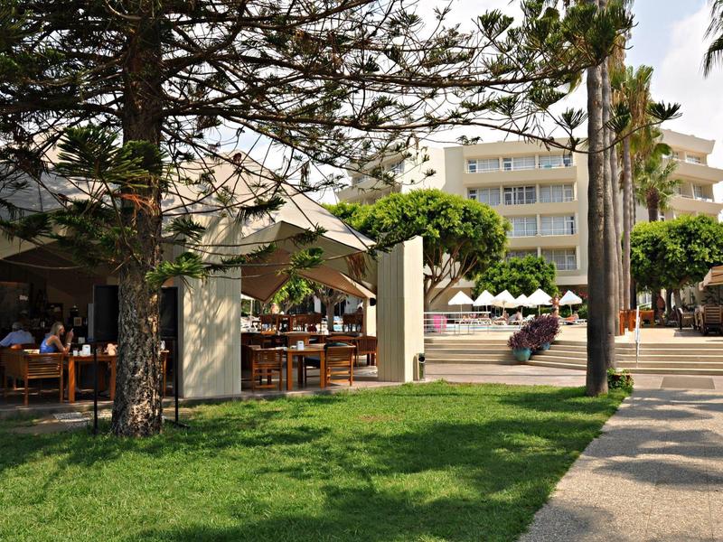 Grüner Park mit Sitzbereichen und Sonnenschirmen vor Gebäuden an einem sonnigen Tag.