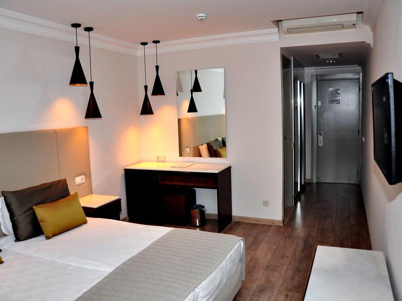 Hell eingerichtetes Hotelzimmer mit Doppelbett, schwarzem Schreibtisch und modernem Fernseher an der Wand.