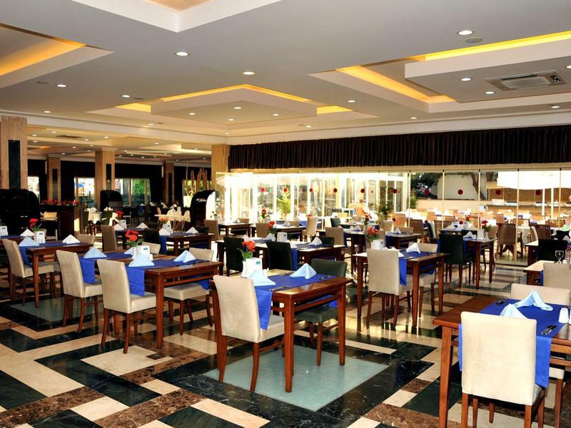 Großer Speisesaal mit Holztischen, weißen Stühlen, blauen Servietten und gemustertem Boden.