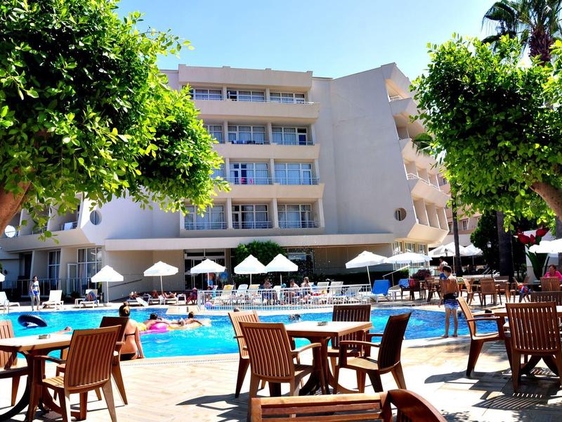 Hotel mit großem Pool, Sonnenschirmen, Holzstühlen und Tischen, umgeben von grünen Bäumen.