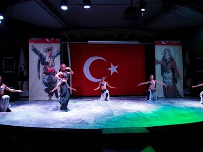 Bambini ballano su un palco davanti a una grande bandiera turca in una stanza poco illuminata.