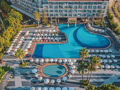 Großer Hotelpool mit Sonnenliegen, Palmen und angrenzendem Hotelgebäude bei klarem Wetter.