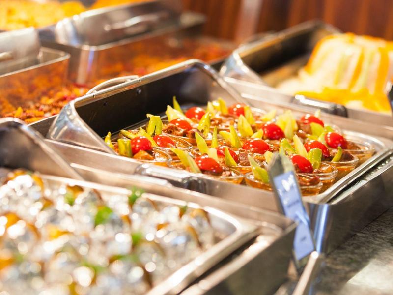 Bufet s různými teplými pokrmy v nerezových nádobách, včetně těstovin s rajčaty a zeleninou.