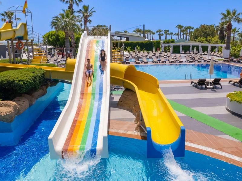 Dva žluté vodní skluzavky vedle bazénu v tropickém letovisku s palmami a lehátky.