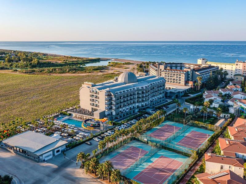 Pohled na hotel s tenisovými kurty blízko moře a otevřenými poli pod jasnou oblohou.