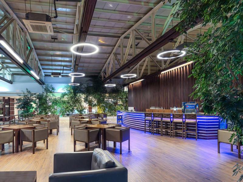 Moderní hotelová restaurace s dřevěnými podlahami, jídelními stoly a osvětleným barem v teplé atmosféře.