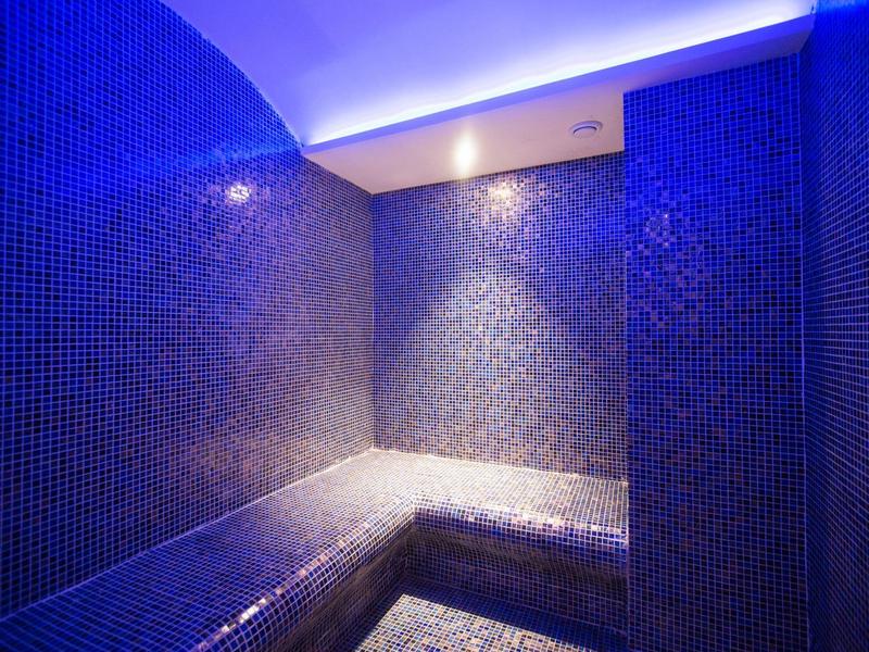 Tmavá moderní parní místnost s modrými mozaikovými dlaždicemi a nepřímým osvětlením stropu.