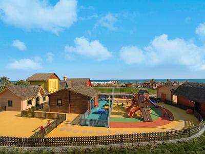 Kleurige vakantiehuizen met speelplaats en zwembad dicht bij het strand onder een blauwe lucht.