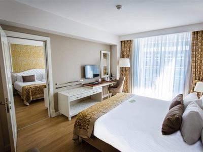 Lichte hotelkamer met houten vloer, wit bed, bureau, tv en deur naar slaapkamer.