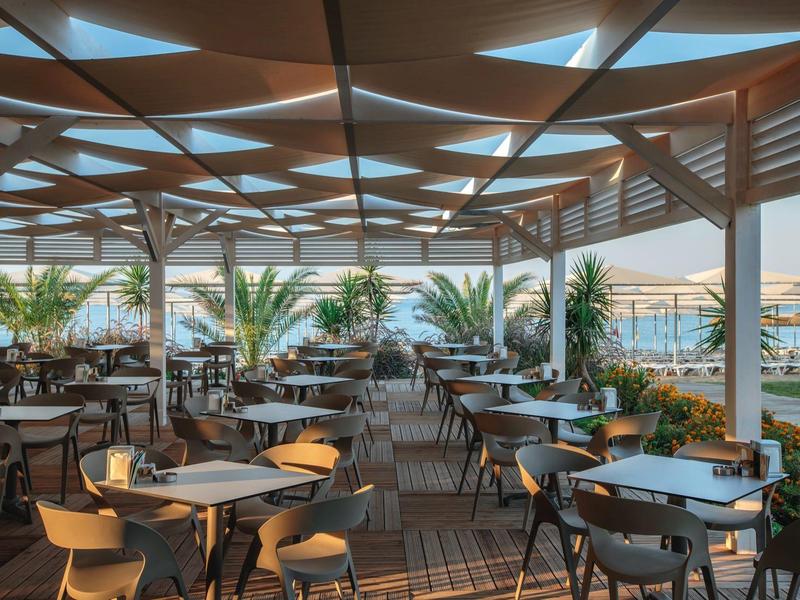 Zastřešená terasa hotelu s stoly, židlemi a výhledem na pláž a palmy.