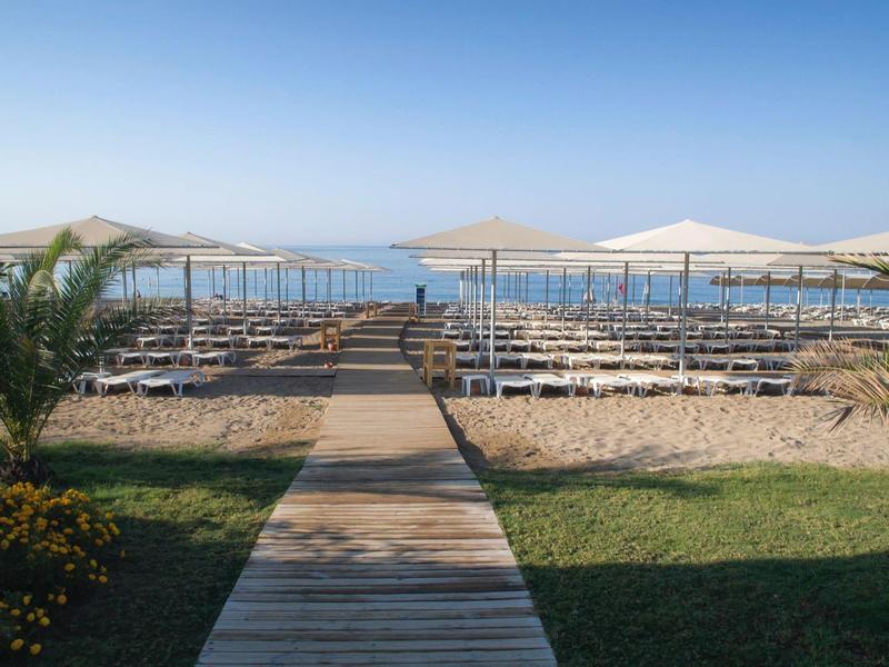 Dřevěná chodníková cesta vede k lehátkům a slunečníkům na pláži s výhledem na moře.