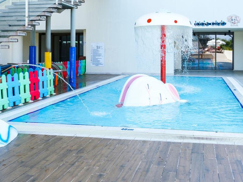 Malý krytý bazén s barevným plotem a vodní hrou pro děti v hotelovém prostředí.