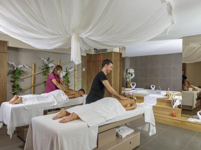 Dva maséři ošetřují klienty v jasné spa místnosti s bílými závěsy a dřevěnými prvky.