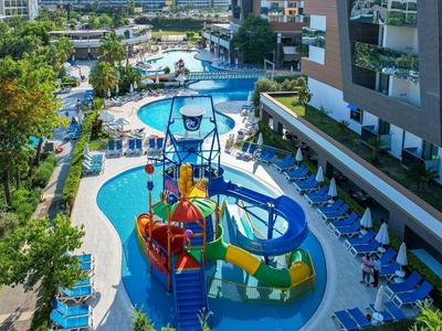 Großer Außenpool mit Wasserrutsche, Spielturm, Liegestühlen und umliegenden Gebäuden an sonnigem Tag.