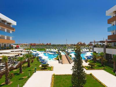Blick auf einen Hotelpool zwischen weißen Gebäuden mit Palmen und blauen Liegen bei blauem Himmel.