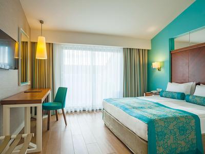 Modernes Hotelzimmer mit Bett, Schreibtisch, Stühlen und großer Fensterfront mit Vorhängen.