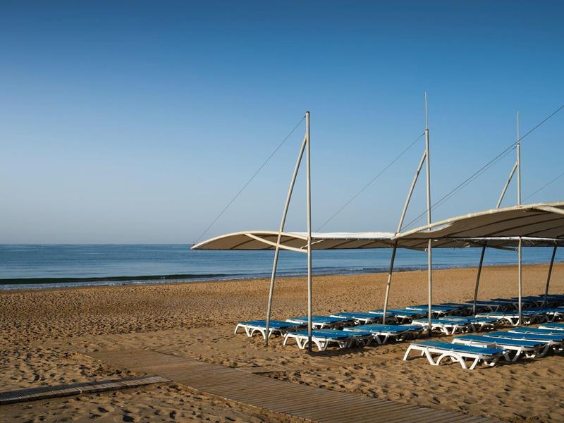 Klidná pláž s modrými lehátky a bílými slunečníky u moře pod jasnou oblohou.