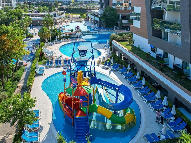 Hotelový bazén s barevnými skluzavkami a modrými slunečníky pod jasnou oblohou.