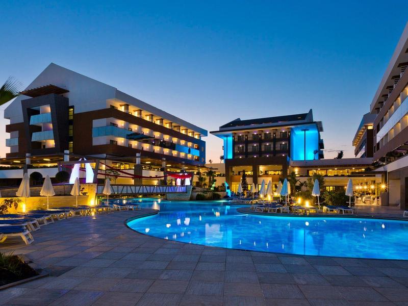 Moderní hotel s osvětleným bazénem a palmami při západu slunce.