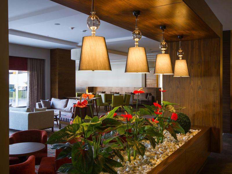 Světlý hotelový salonek s závěsnými lampami, rostlinami a pohodlnými sedadly.