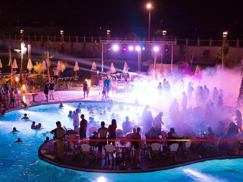 Noční bazén s barevnými světly a mlhou, veselá atmosféra a dav lidí.