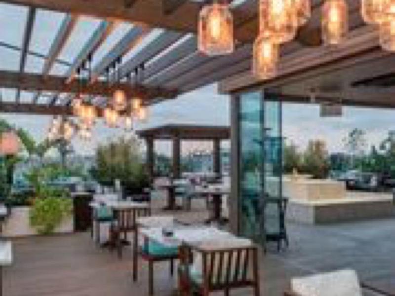 Moderní venkovní prostor hotelu s dřevěnou podlahou, skleněnou střechou a stylovým osvětlením.