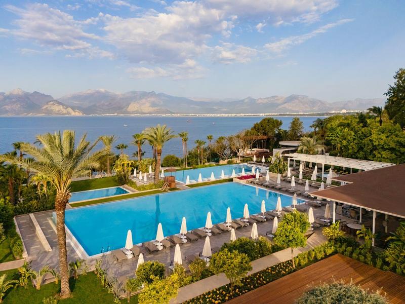 Velký venkovní bazén s lehátky, palmami a výhledem na moře a hory.