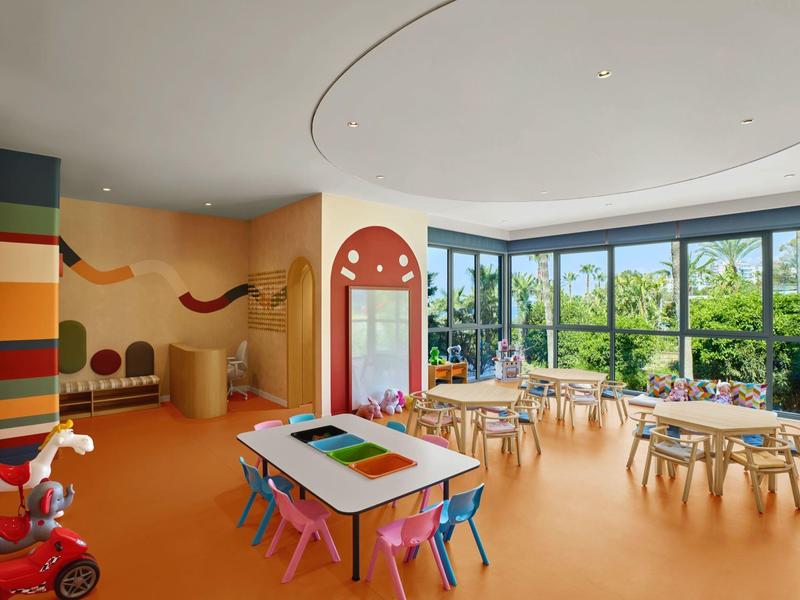 Světlá a barevná dětská herna s stoly, židlemi a velkými okny.