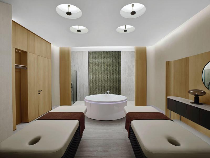 Moderní spa místnost s volně stojící vanou, dvěma lehátky a minimalistickým dřevěným nábytkem.