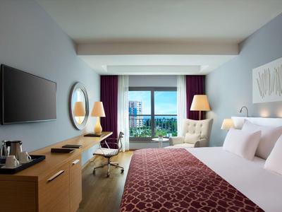 Modern hotel room with large bed, desk, and sea view through a window.