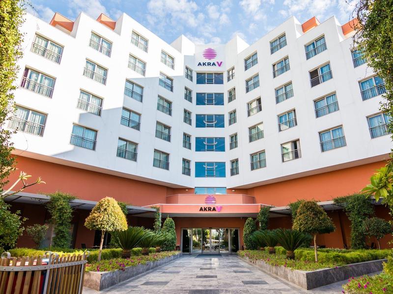 Ala de hotel moderna blanca con entrada roja, rodeada de árboles verdes y cielo azul.