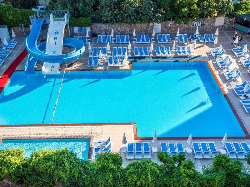 Grande piscine extérieure avec toboggan aquatique, chaises longues bleues et parasols blancs.