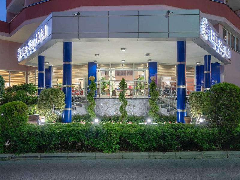 Entrée élégante d'un hôtel éclairé entouré de buissons verts et de colonnes bleues.