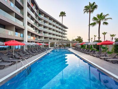 Moderner Hotelpool mit Liegestühlen und roten Sonnenschirmen, umgeben von Palmen bei Sonnenuntergang.