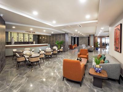 Moderní hotelový salonek se šedými dlaždicemi, oranžovými křesly a jídelními stoly.