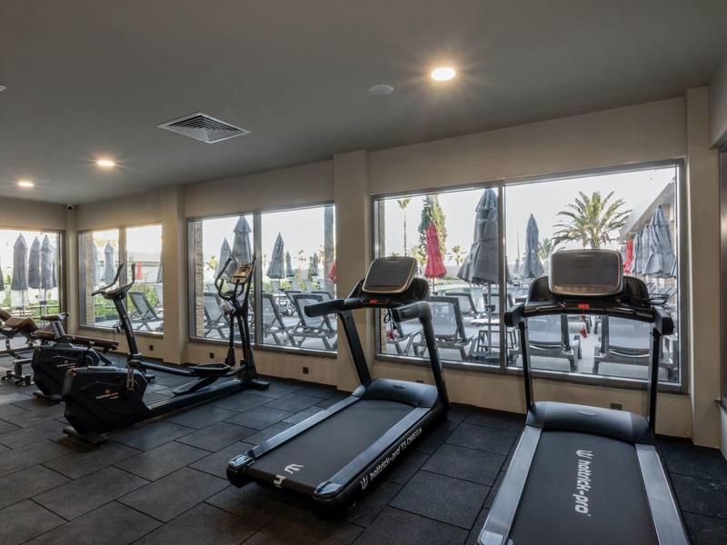 Interiér posilovny s běžeckými pásy s výhledem na venkovní bazén hotelu.