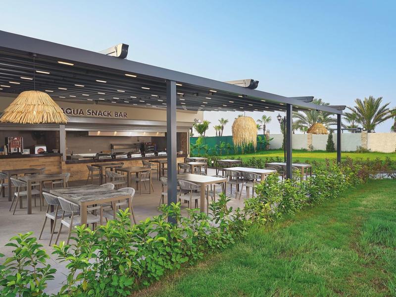 Venkovní restaurace s stolky a židlemi obklopená zelení pod modrou oblohou.