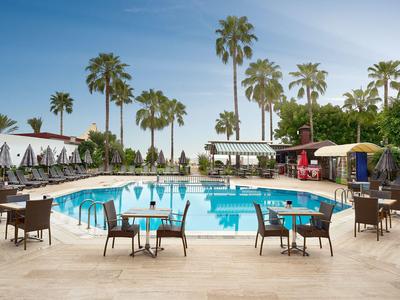 Ein Außenpool mit Tischen, Stühlen und Sonnenschirmen, umgeben von Palmen unter blauem Himmel.