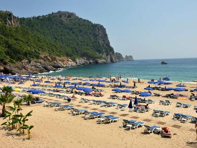 Spiaggia con molte sdraio e ombrelloni blu davanti a una collina boscosa e il mare.