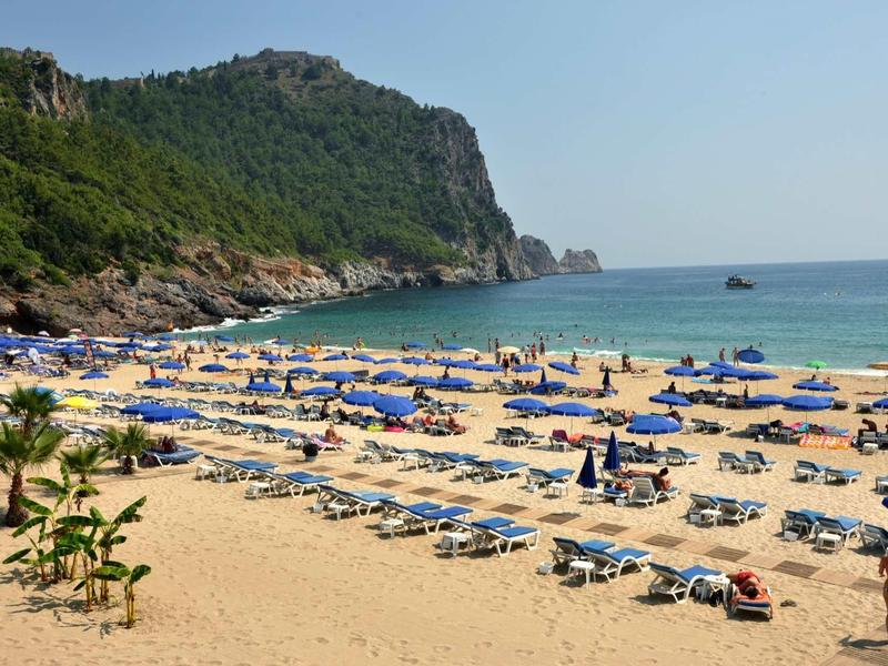 Ein Sandstrand mit vielen blauen Liegestühlen und Sonnenschirmen, dahinter grüner bewaldeter Hügel.