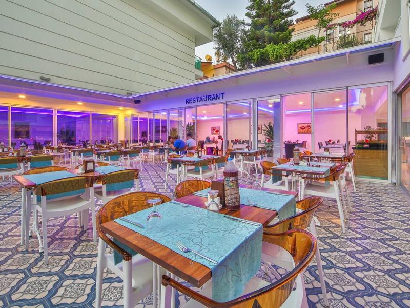 Zweistöckiges Café mit blau beleuchteter Terrasse, Holztischen und Stühlen auf gemustertem Boden.