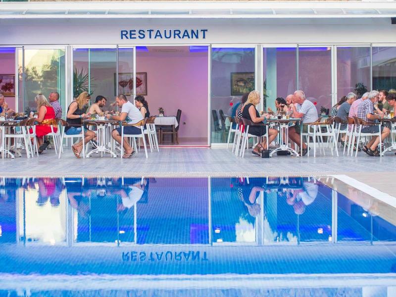 Ein Restaurant mit Gästen an Tischen neben einem spiegelglatten blauen Pool unter hellem Licht.