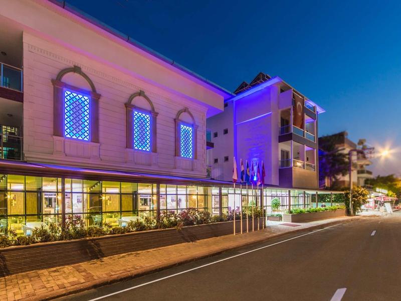 Beleuchtetes, mehrstöckiges Gebäude mit bunten Fenstern und angrenzender Straße bei Nacht.