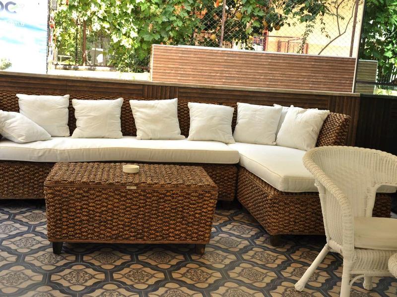 Gemütliche Ecklounge aus braunem Rattan mit weißen Kissen und passendem Tisch auf gemustertem Boden.