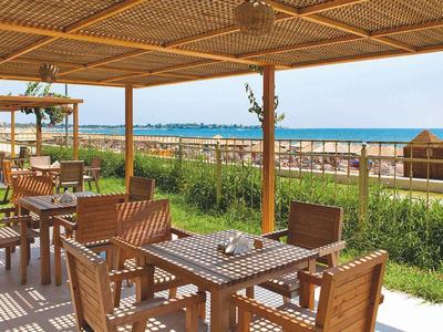 Offenes Café mit Holztischen und Stühlen, Blick aufs Meer, grüner Rasen und Strand im Hintergrund.